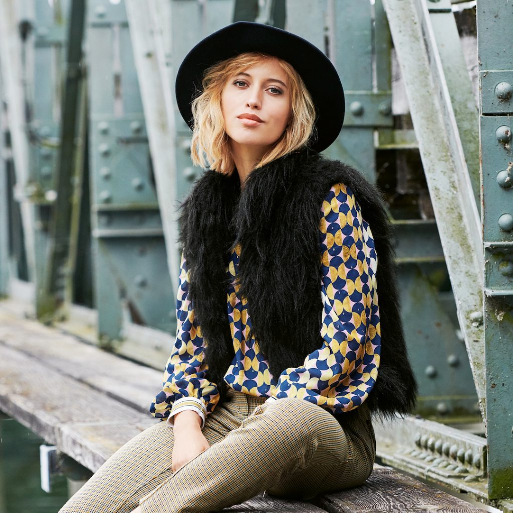 Model wearing a black hat, the black waistcoat out of long fake fur over a blue and yellow blouse and beige trousers.