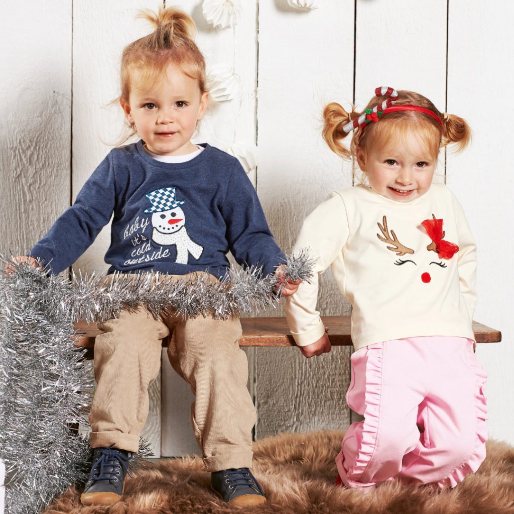 A boy and a girl are shown, the boy is sitting on a bench and wears a blue sweater and brown cord trousers, the girl is leaning on the bench while smiling, wearing two pigtails, a white sweater with a reindeer motive and the pink "Candy" trousers with frills on the side, running from the waist to the hem of the trousers.