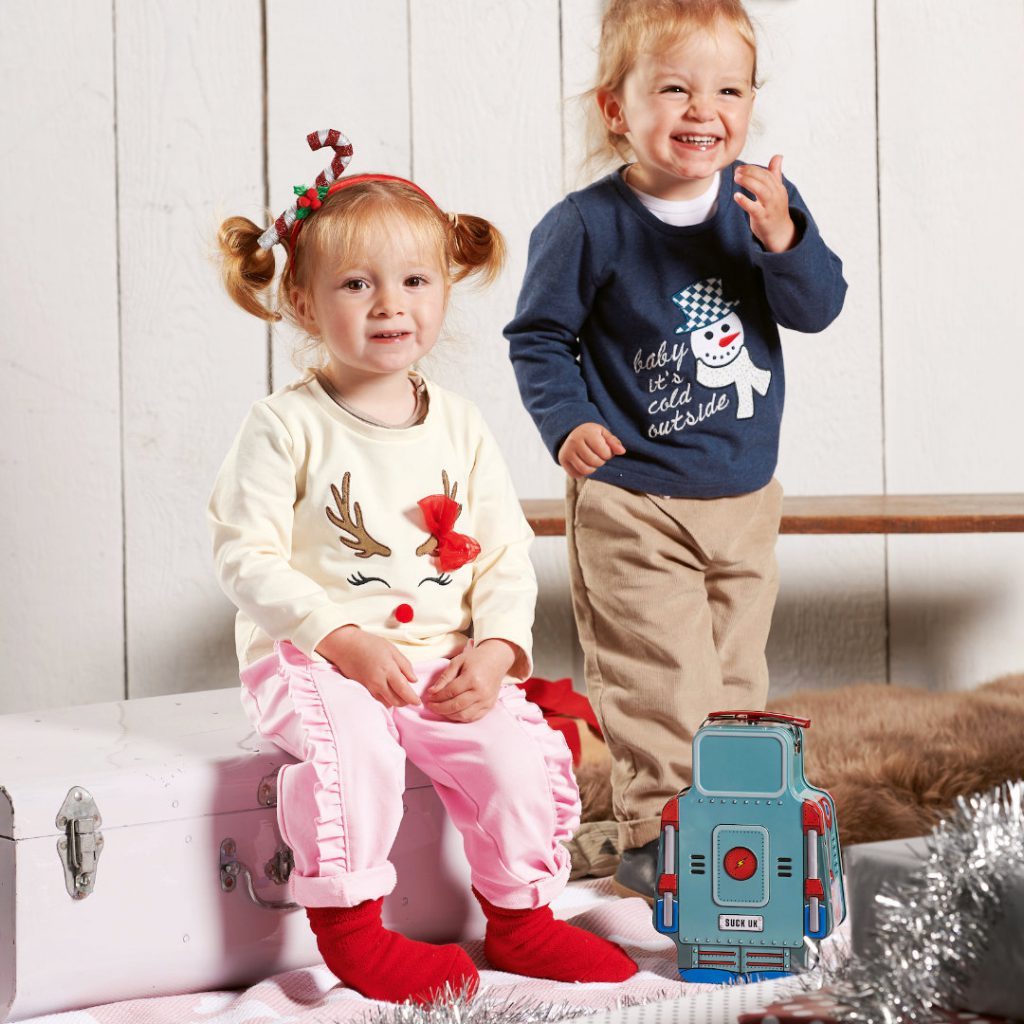 A boy and a girl are shown, the boy is standing and laughing, with his left hand near his mouth, his chin has spots from chocolate on it. He wears a blue sweater and brown cord trousers. In front of him stand a robot toy. The girl is sitting on a pink suitcase to the right of the boy, wearing two pigtails, a white sweater with a reindeer motive and the pink "Candy" trousers with frills on the side, running from the waist to the hem of the trousers. She has red socks.