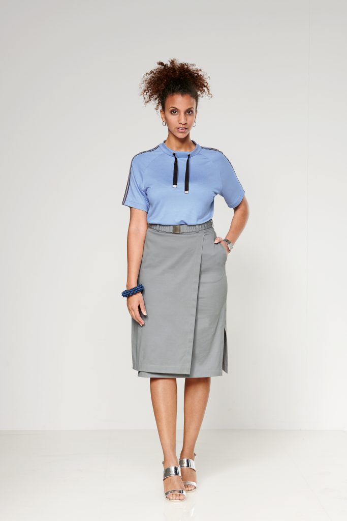 Model wearing a skyblue tshirt with sleeves reaching the hight of her ellbows, with a silver stripe fom ellbow to shoulder. The Shirt also has a black cord around the neck. The shirt is tucked in a beige skirt called "Malu".