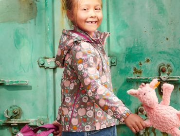 a child with ponytail is walking in front of a turqoise container door, wearing the coat made out of a patterned fabric in brown, pink, green and orange, also wearing a jeansdress. She has a pink school backpack in the left hand and a blush pink sheep plush in the right hand. She is smiling and one of her front teeth is missing.