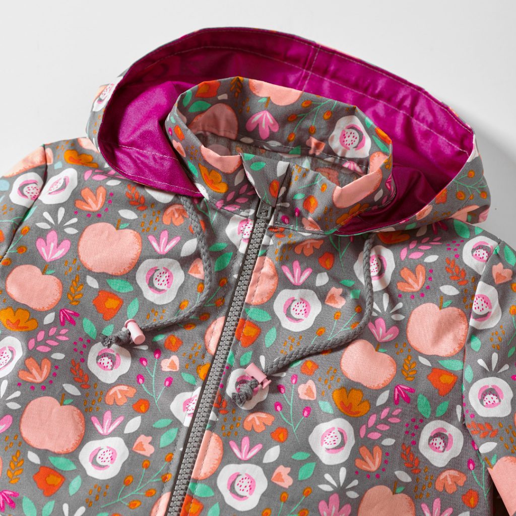 A close up of this month's free sewing pattern, a brown-greyish coat with pink apples, green leaves and orange flowers as a pattern. The Hood with hot pink inside has two grey cords and blush pink cord stoppers, so the hood can be tied and stays up in bad weather. The grey zipper is closed and has a little triangle fabric piece at the top so it does not rub against the chin and protects the skin.