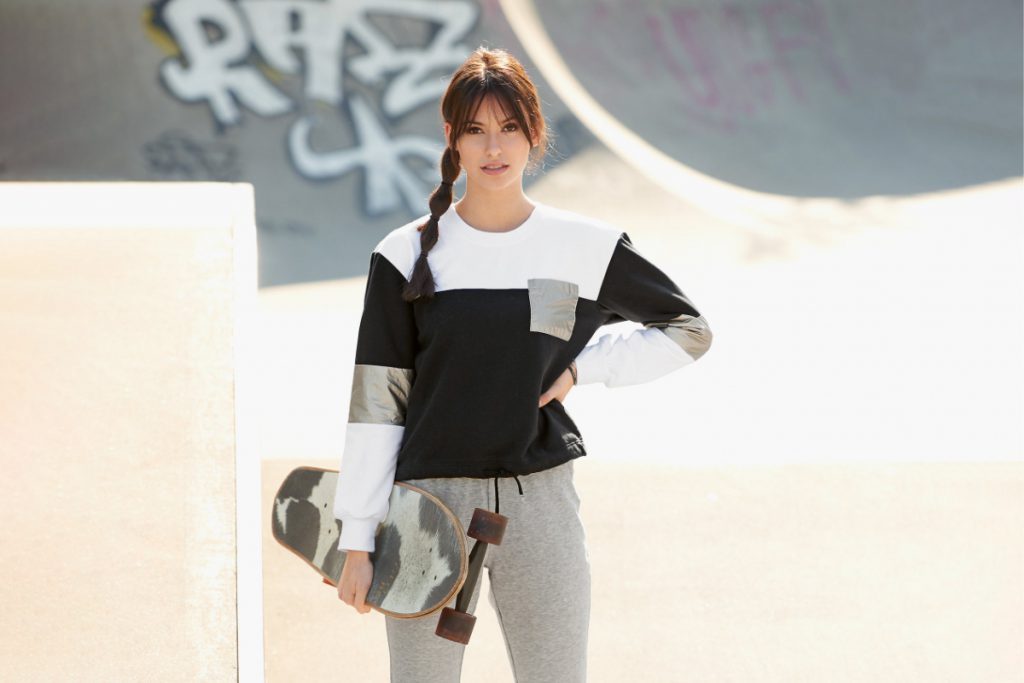 A woman in a skatepark carries a used looking skateboard in her right hand. Her left hand ist on her waist. She wears the free pattern of december, the sweater Luna with white, black and silver fabrics and grey yoga pants.