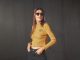 The shirt "Vio" is mustard yellow and has wavy rolled hems on the sleeves, waist and neckline. It is made out or ripped jersey and features a black embroidered design saying "Weekend, I'm ready for you". The brown haired model with long hair wears sunglasses and black jeans with the crop top.