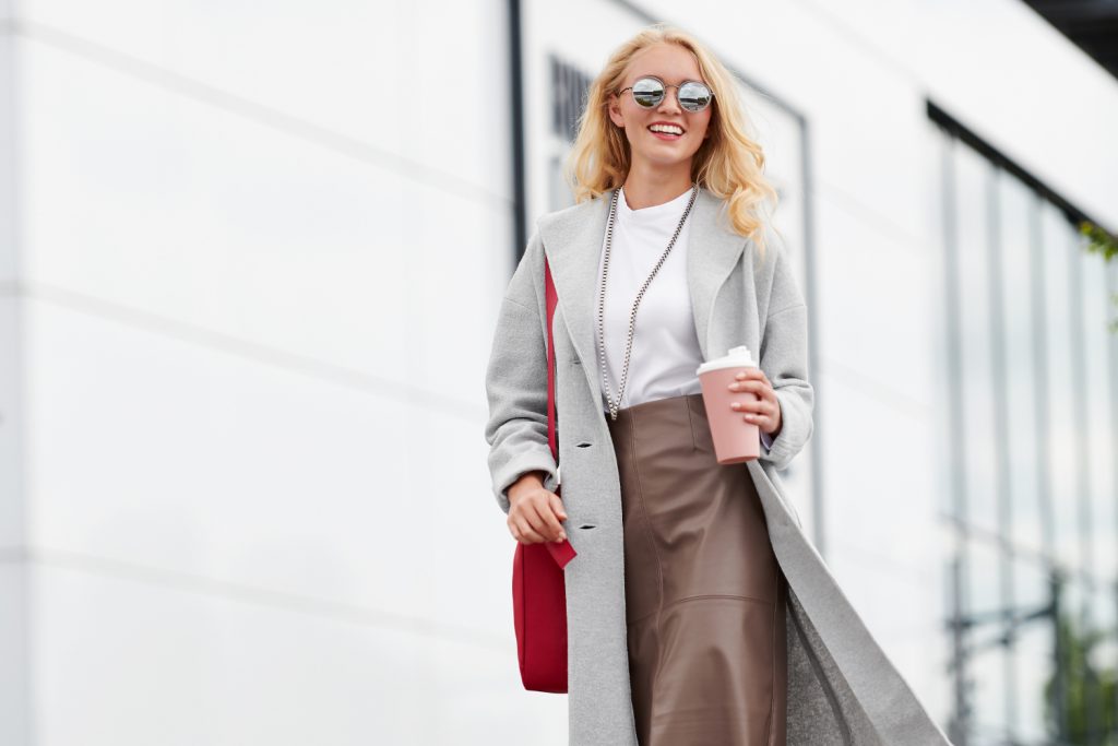 A model with long, blond hair is walking in front of a white building, wearing a grey felt coat, a brown faux leather skirt and white t-shirt. She has a long silver chain around her neck and wears mirrored, round sun glasses. Her left hand carries a pink coffe cup, and she has the free pattern of the month, the handbag "Klara" on her right shoulder.