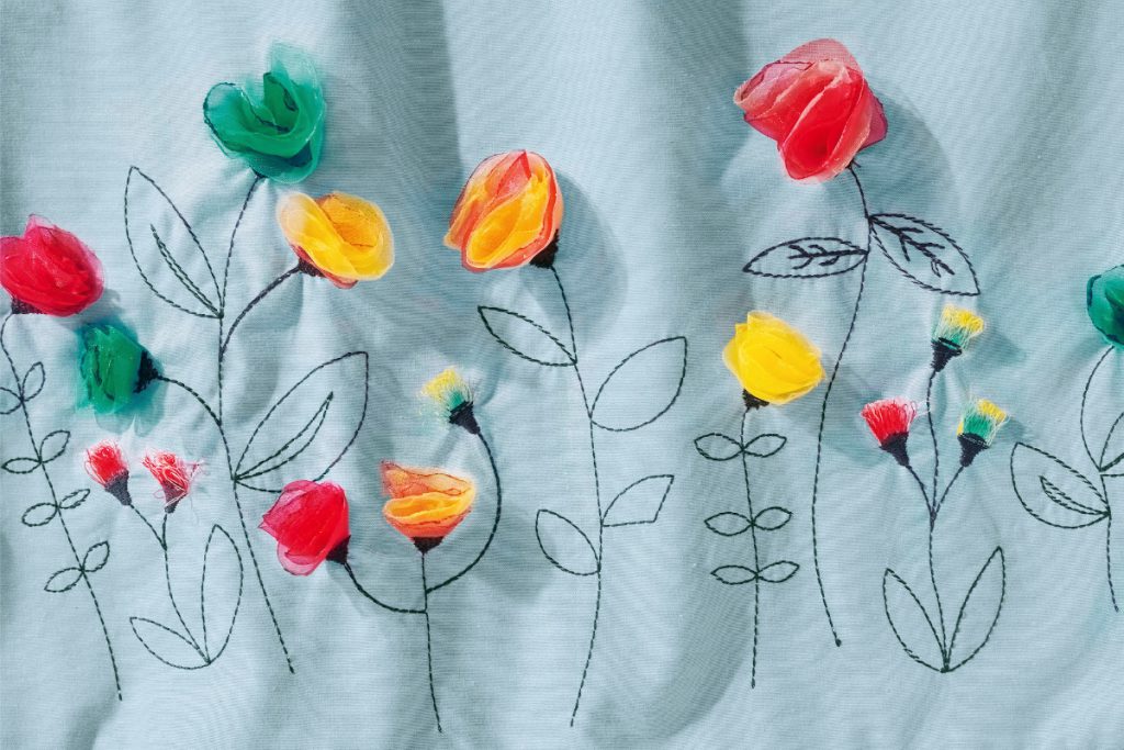 Detailed view of the 3D flower embroidery of this month's free pattern. The flower buds in various colors are attached to stems with leaves, stitched as black lines.