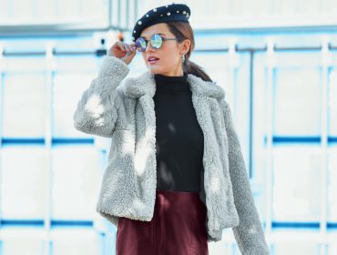 The blueish-grey jacket made from fluffy teddy fabric has a straight cut and a wide collar. It is styled with a black turtle-neck shirt, a bordeaux faux leather skirt, round sunglasses and a beret with attached pearls.