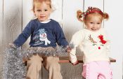Two toddlers wear sweaters with applique designs. The boy on the left wears a blue sweater with a snowman, and the girl on the right a beige one with a reindeer. The embroidery designs are part of the free sewing pattern of this month